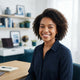 A diverse woman smiling in a bright, modern home office setting.